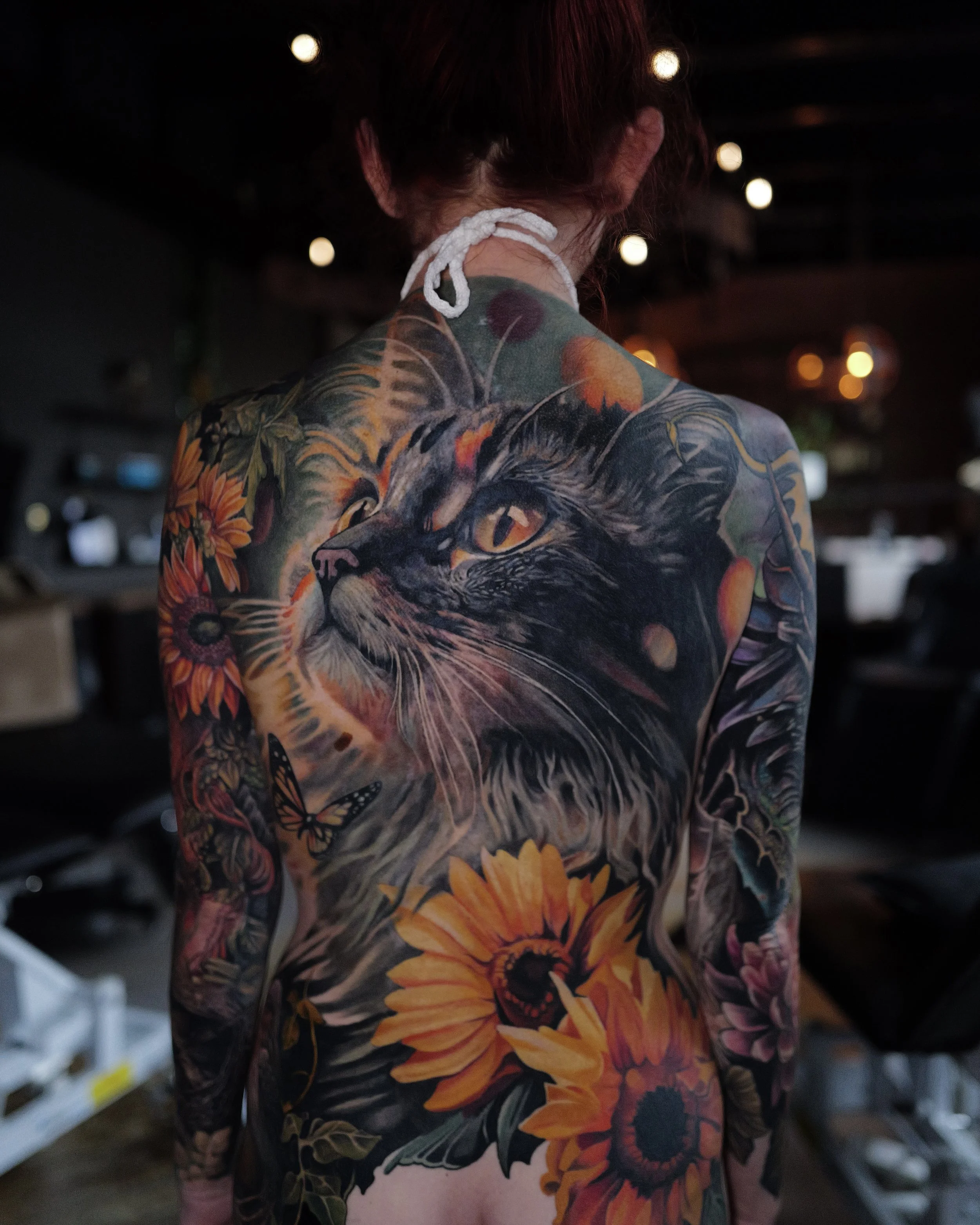 A woman with a large colorful tattoo of a cat surrounded by sunflowers, butterflies, and other floral elements covering her back.