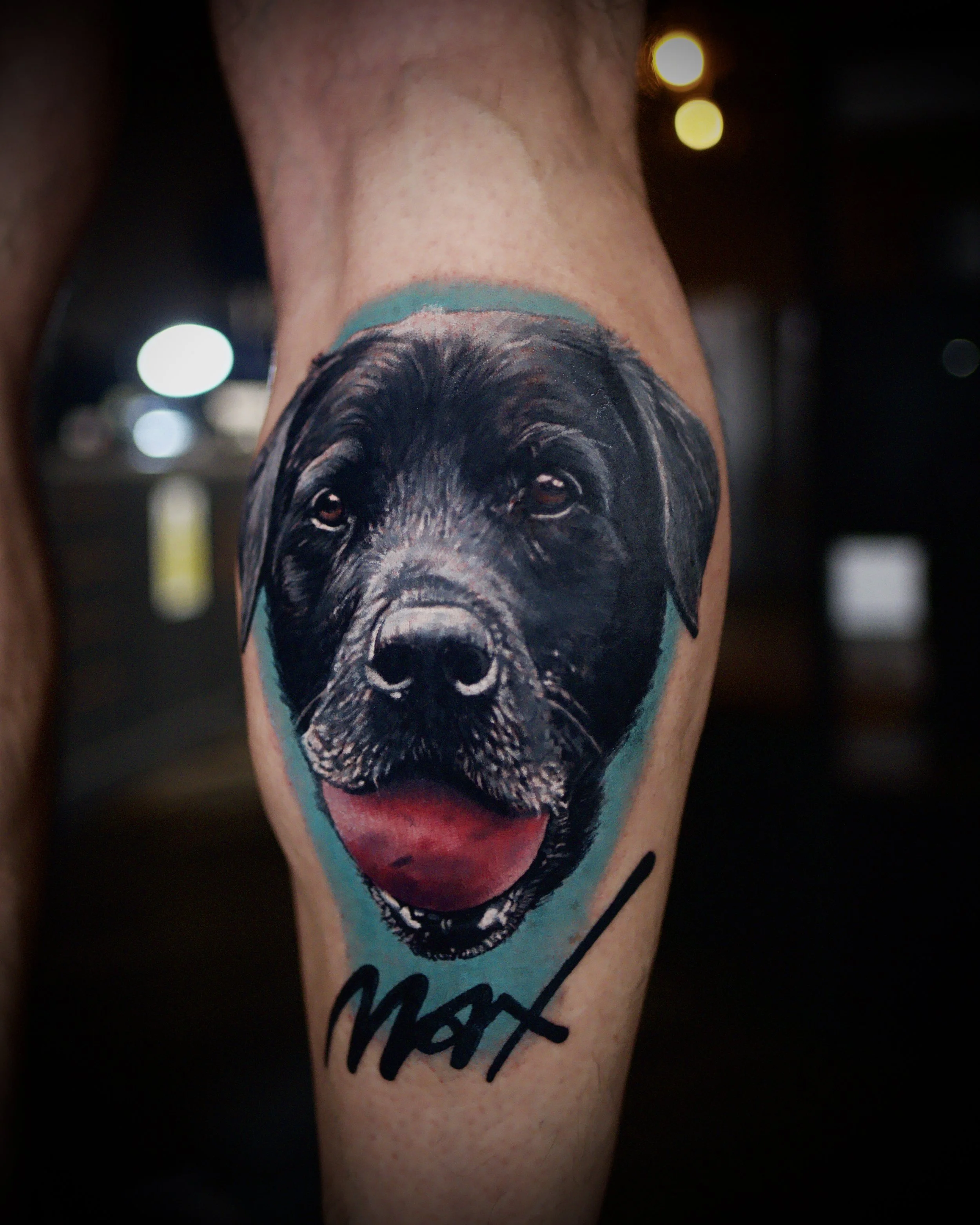 Tattoo of a black Labrador retriever's face with a pink tongue and brown eyes on a person's leg, with the name 'Max' written below.
