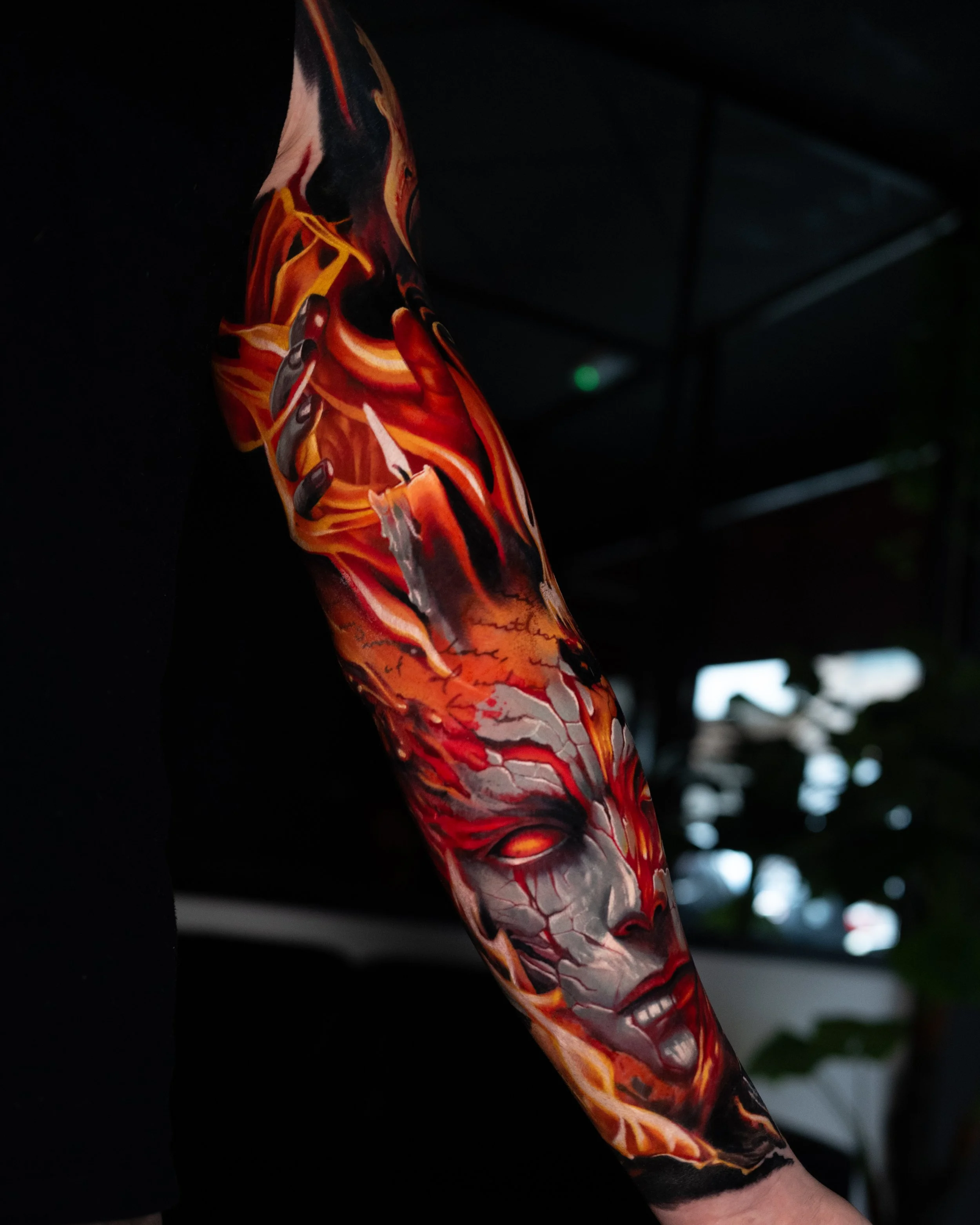 Colour realism tattoo by a tattoo artist from Manchester - Tibor Varga. Close-up of a colorful, fiery tattoo sleeve on a person's arm, featuring a woman's face with gray cracked skin and glowing eyes surrounded by flames and abstract designs.