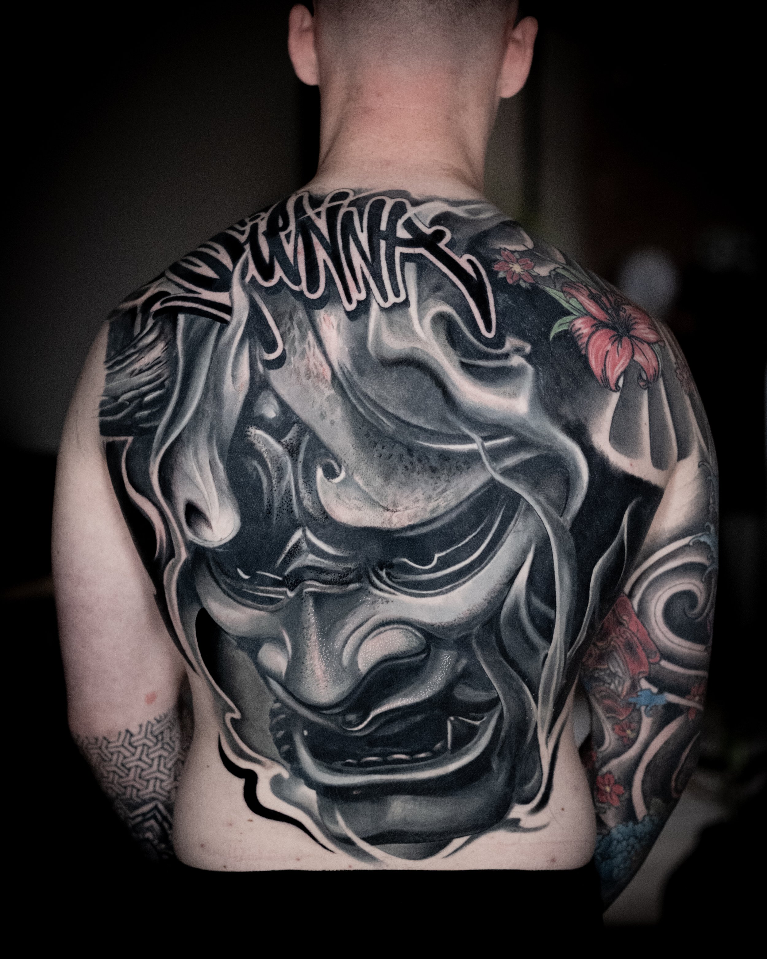 Back tattoo of a fierce traditional-style demon or ogre face with a wide mouth, sharp teeth, and intense expression in black and gray ink, with colorful flower tattoos on the shoulders.