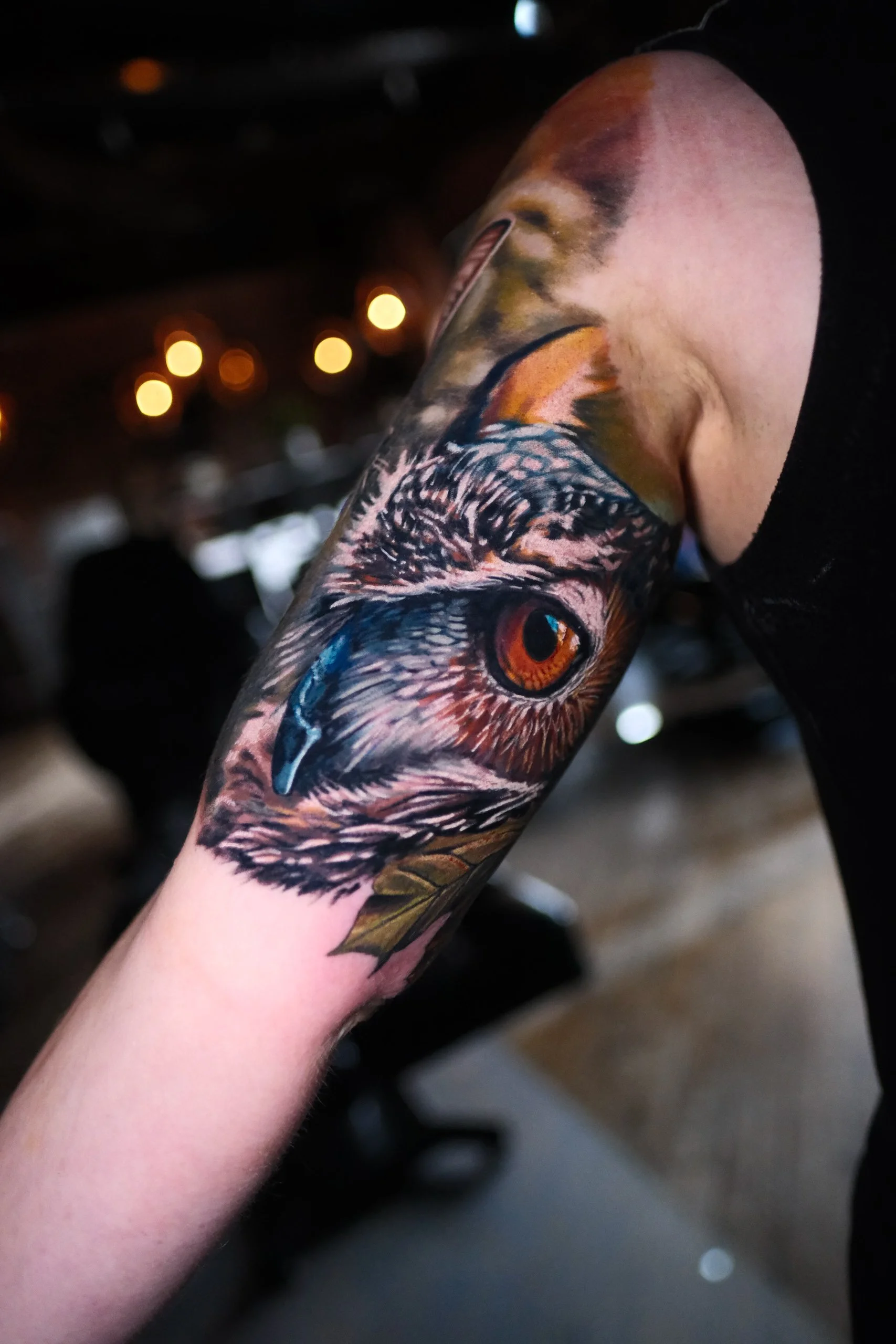 Close-up of a person's arm with a detailed tattoo of an owl's face and eye, featuring orange and black colors.