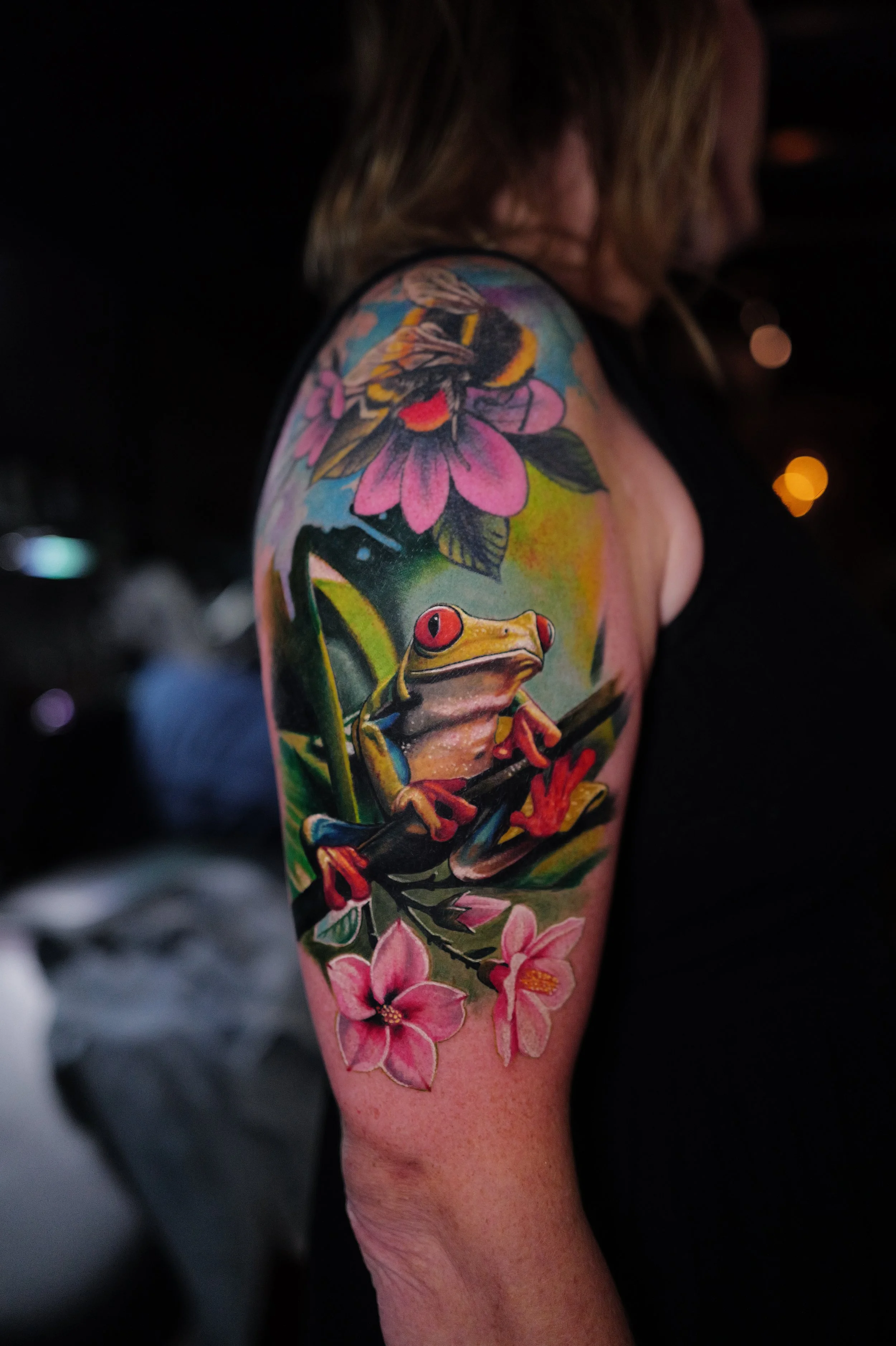 Colorful tattoo on woman's upper arm featuring a frog sitting on a branch, surrounded by pink flowers and a bee.
