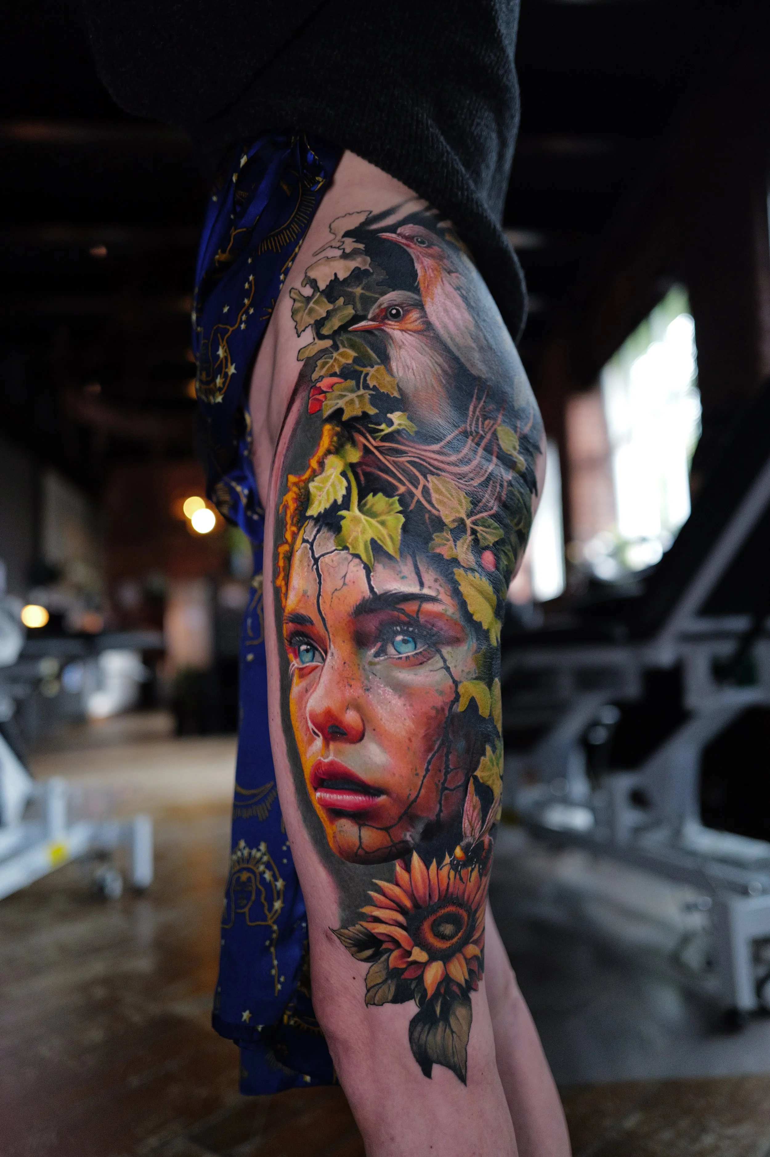 Colorful tattoo of a woman's face with blue eyes, surrounded by leaves, birds, a sunflower, and a moth on a person's leg.