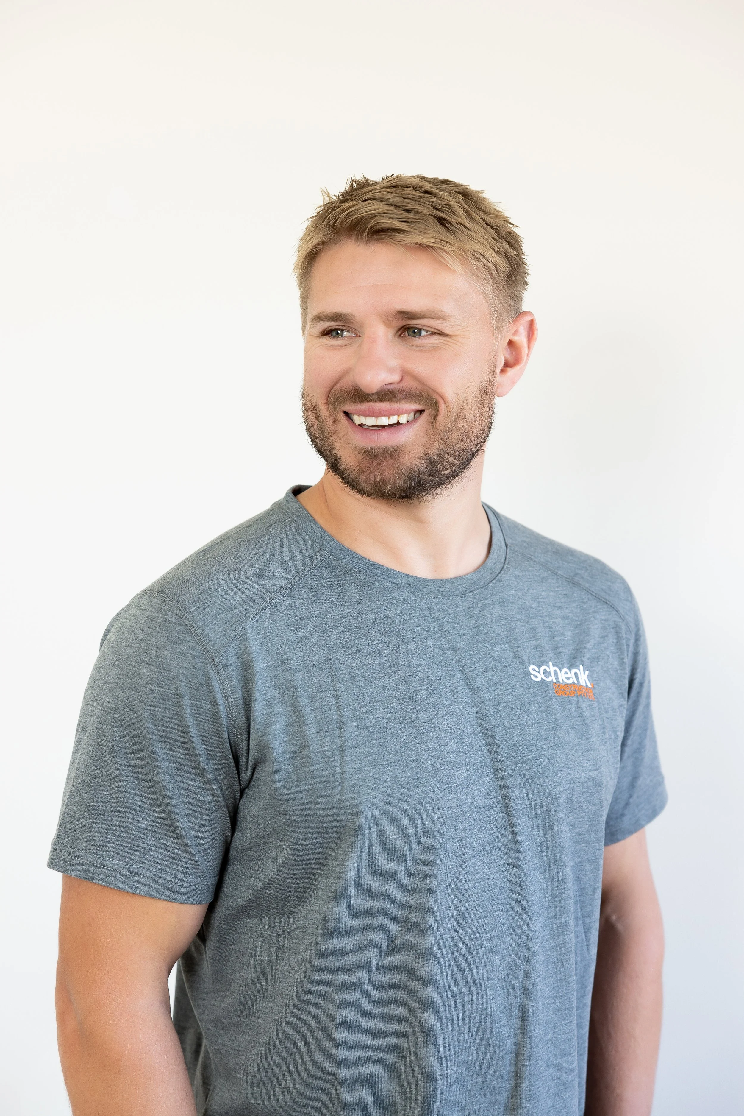 A smiling man with short blond hair and a beard, wearing a gray t-shirt with 'Schenk' logo on the chest, standing against a white background.