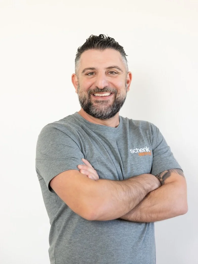 A smiling man with dark hair, beard, and mustache, wearing a gray t-shirt with 'schenk' on it, standing with arms crossed against a plain white background.