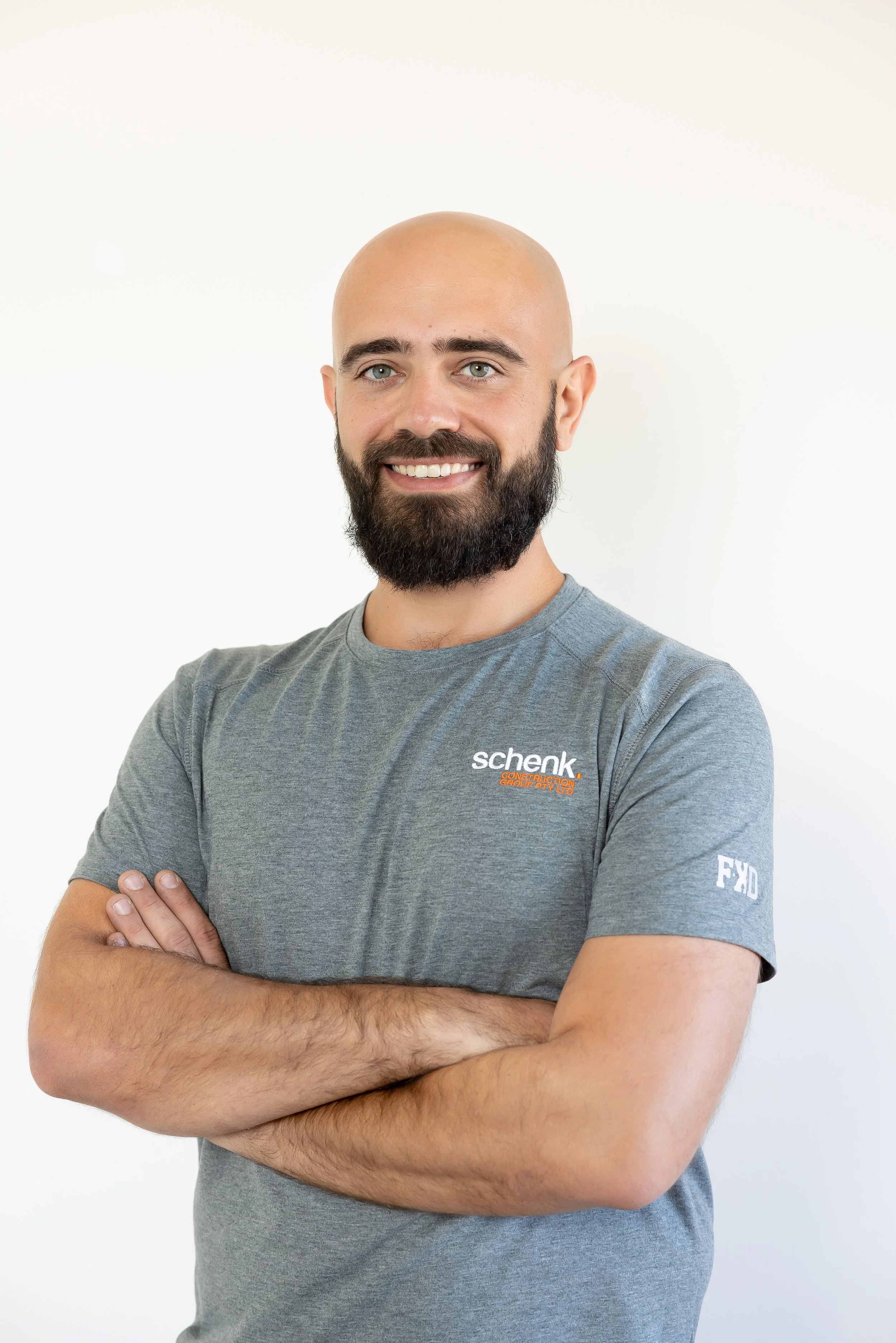 A man with a beard and bald head, smiling, with arms crossed, wearing a gray t-shirt against a plain white background.