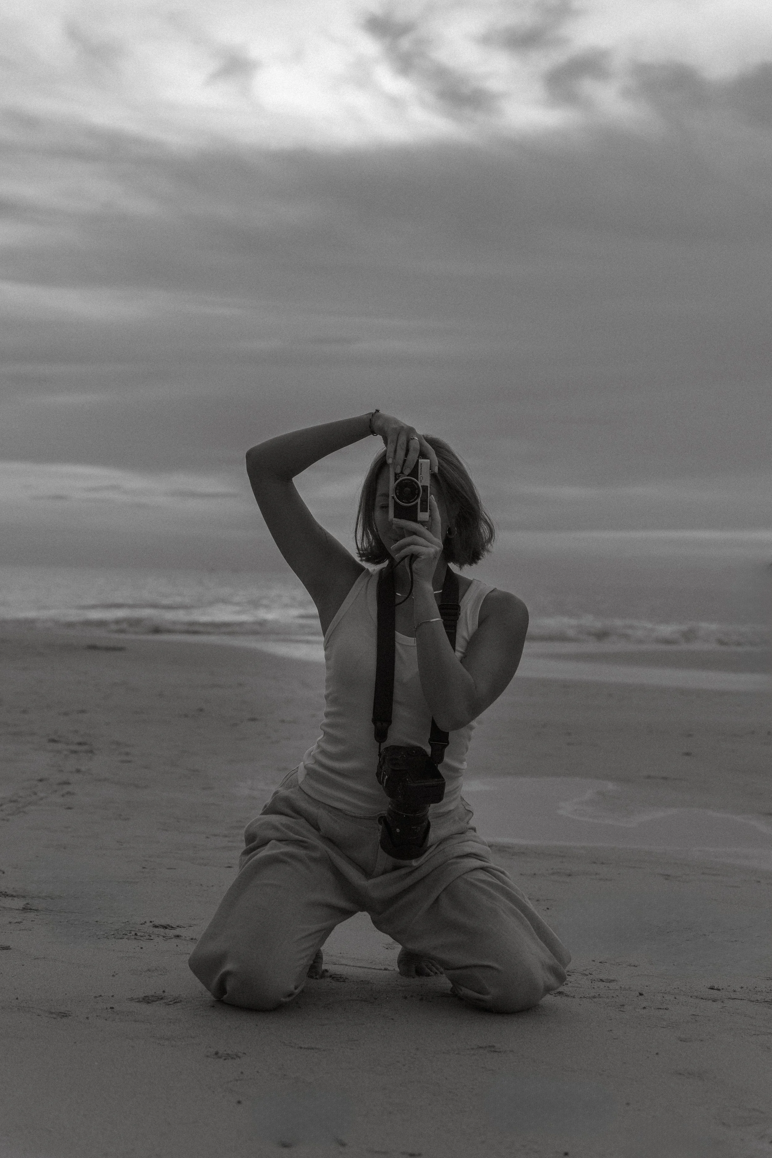 A woman with short hair taking a photograph on a beach during sunset or sunrise, kneeling on the sand with waves in the background.