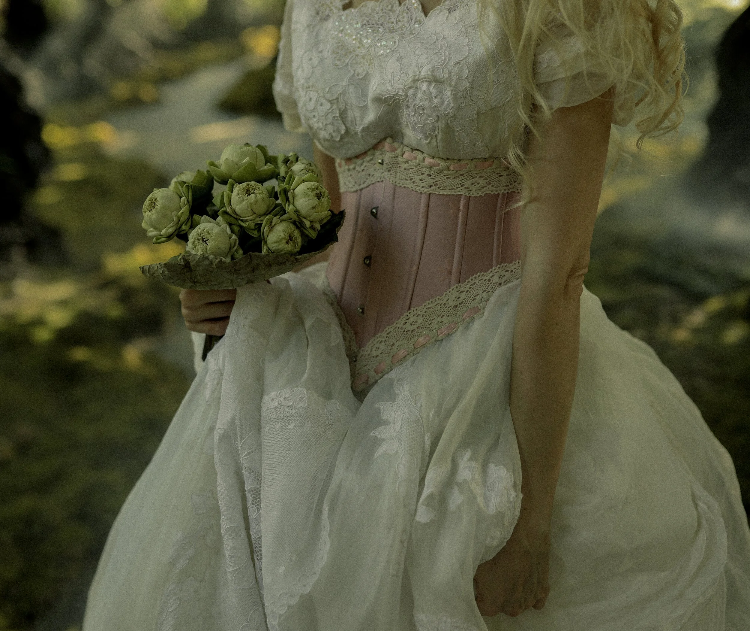 Close-up of a woman wearing a vintage white dress with lace details and a pink corset, holding a bouquet of white peonies, outside in a forested area.