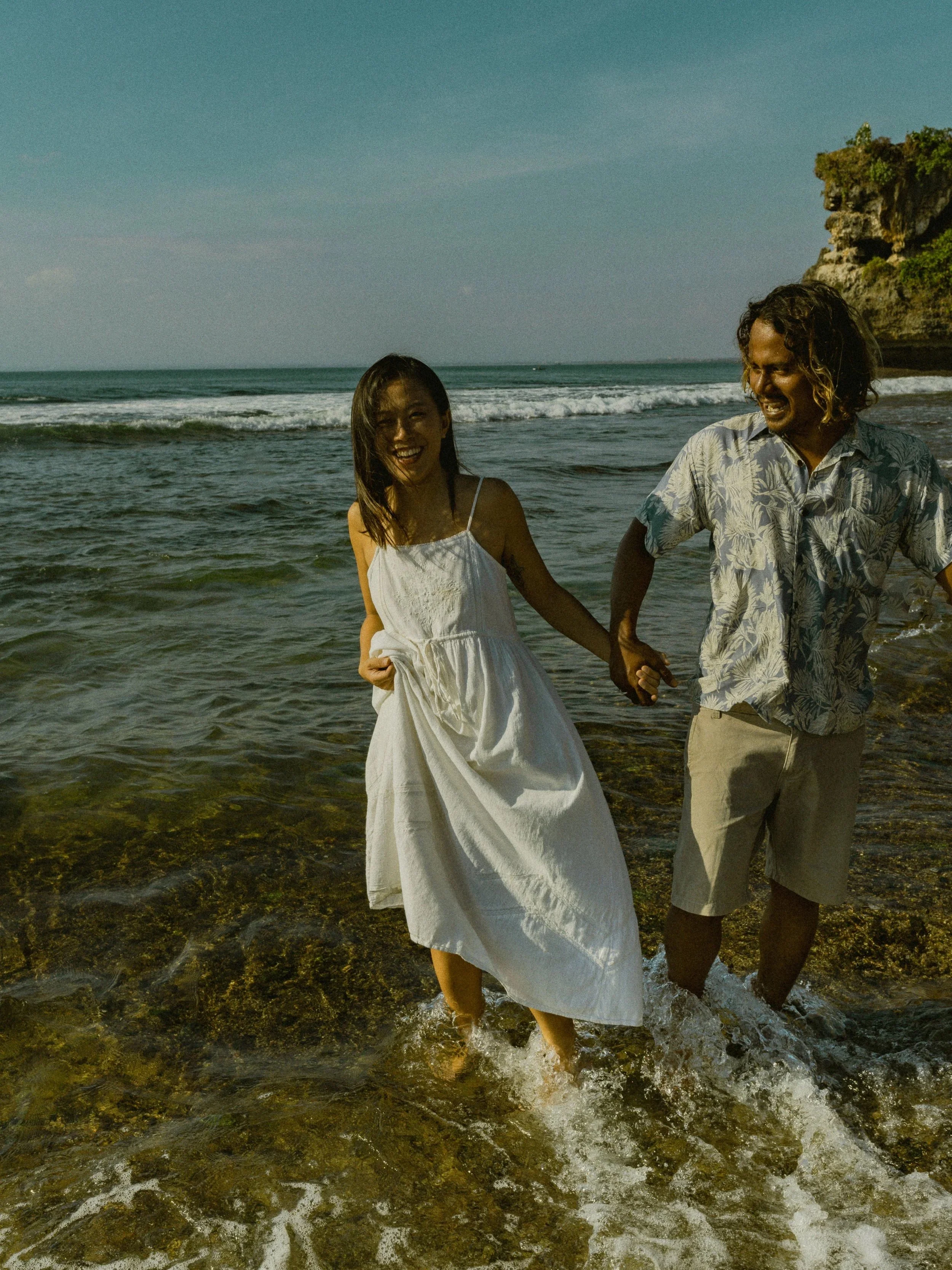 A couple holding hands and smiling in the ocean, wearing casual summer clothes with waves around their feet.