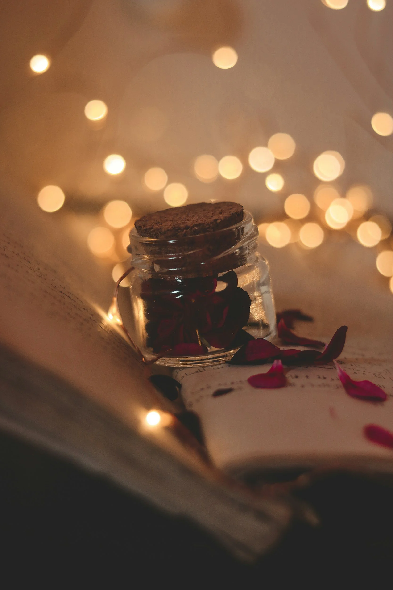 A small glass jar with a cork lid containing dried rose petals, placed on an open book with scattered rose petals around it, illuminated by warm, blurry string lights creating a cozy atmosphere.