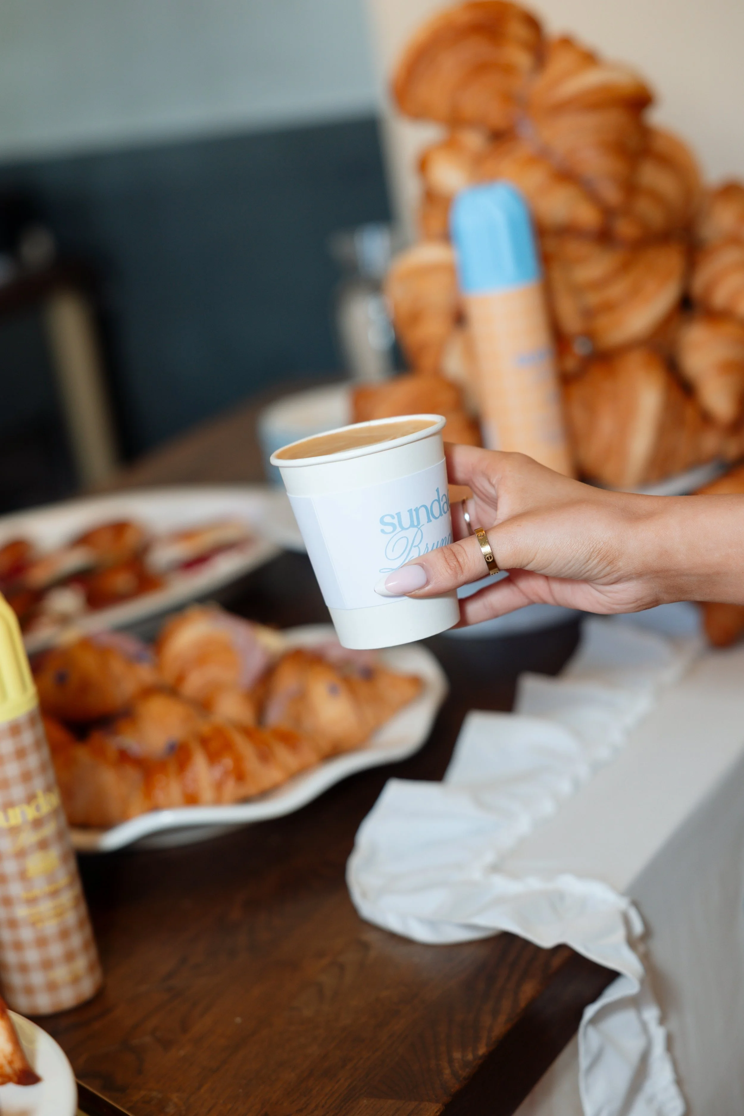 An influencer holding a coffee cup in front of a bakery tablescape at a product launch event for the brand Sundae
