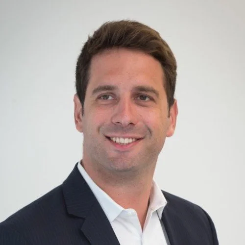 A man with brown hair in a suit smiling against a plain white background.