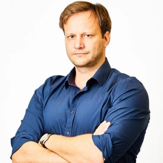A man with light skin, short light brown hair, wearing a dark blue button-down shirt with the sleeves rolled up, crossing his arms and looking serious against a plain white background.