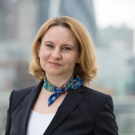 Professional woman with blonde hair wearing a dark blazer and a colorful scarf, standing in an office building with city view.