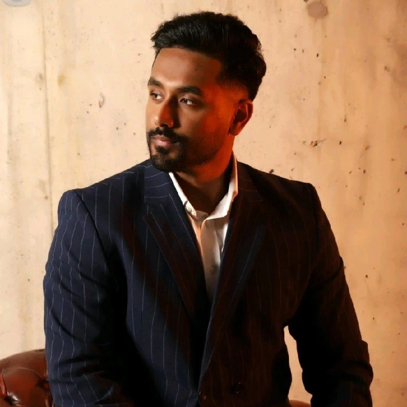 A man with dark hair and a beard, wearing a dark pinstripe suit and white shirt, looking away from the camera in front of a beige wall.