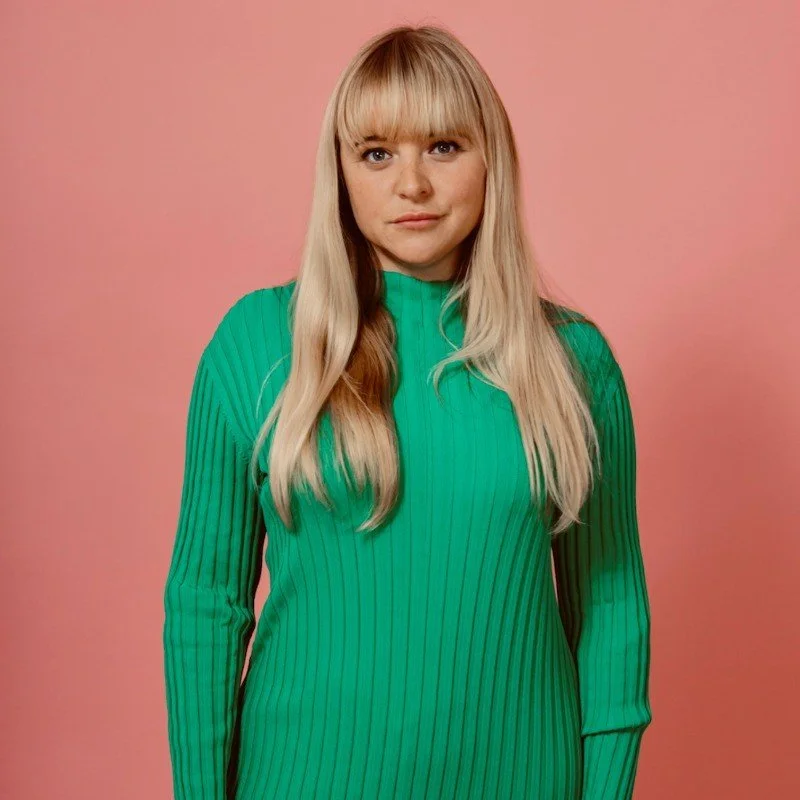 A young woman with long blonde hair and bangs wearing a green ribbed long-sleeve top standing against a pink background.
