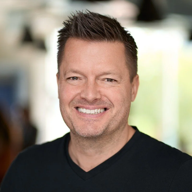 Headshot of a smiling man with short, spiked dark hair wearing a black shirt. Blurred indoor background with natural light.