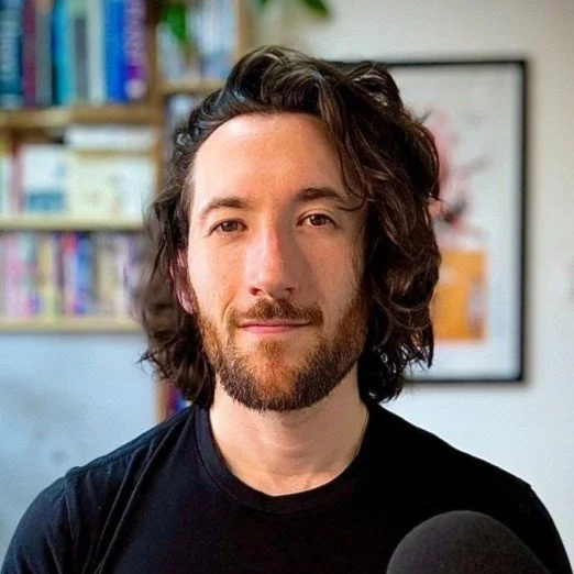 A man with long, wavy brown hair and a beard, wearing a black shirt, sitting indoors with a bookshelf and a framed picture in the background.