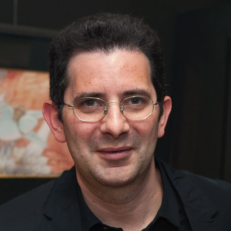 A man with dark hair, glasses, and light skin, wearing a black jacket and shirt, smiling indoors with a blurred artwork in the background.