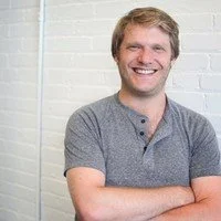 A smiling young man with light brown hair, wearing a gray T-shirt, standing against a white brick wall.