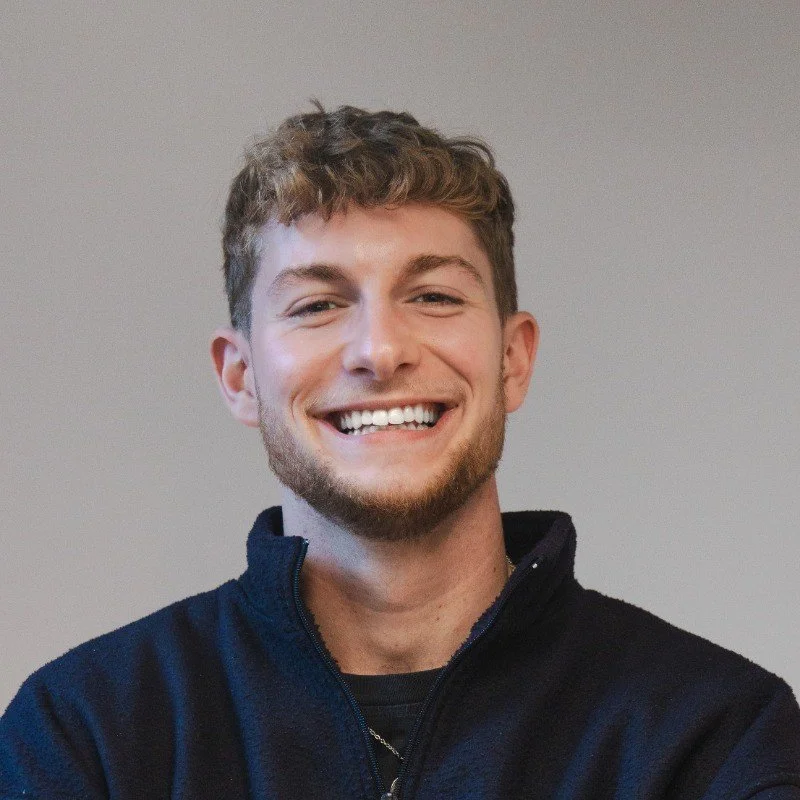 A young man with light brown hair, a beard, and a big smile, wearing a black jacket, standing against a plain background.