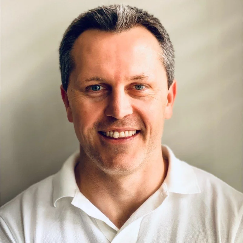 A smiling middle-aged man with short brown hair wearing a white collared shirt.