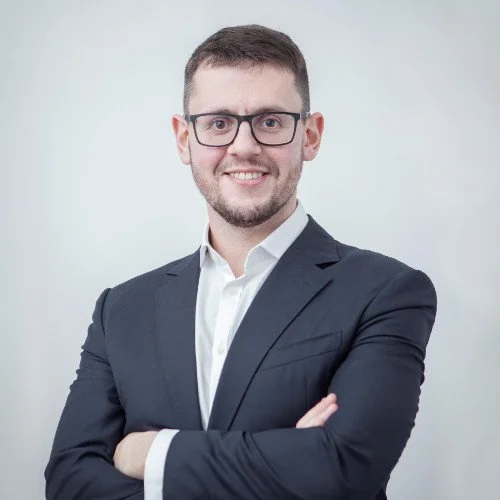 A man in a suit with glasses smiling, standing with arms crossed against a plain light gray background.