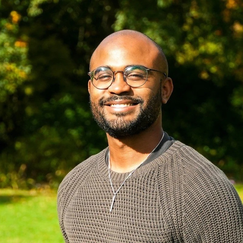 Smiling man outdoors with trees in the background, wearing glasses, a textured sweater, and a necklace.