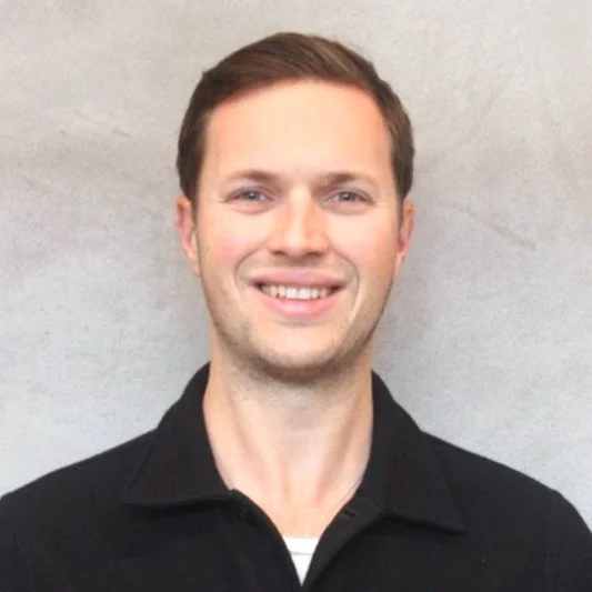 Portrait of a man with brown hair, smiling, wearing a black coat over a striped shirt, standing against a light gray background.