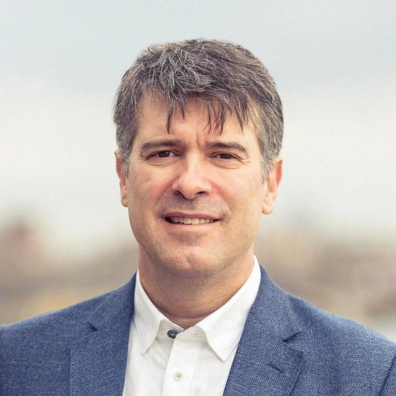 Portrait of a middle-aged man with short brown hair, wearing a white shirt and a dark blue blazer, smiling outdoors with a blurred background.