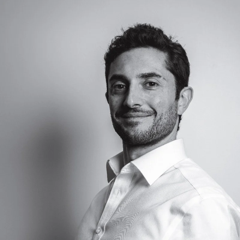Black and white portrait of a man wearing a white collared shirt, smiling lightly with a neutral background.