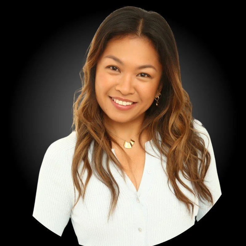 A woman with long wavy brown hair smiling, wearing a white button-up shirt, gold jewelry, against a dark background.