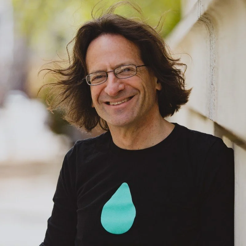 A smiling man with glasses and shoulder-length dark hair, wearing a black T-shirt with a turquoise teardrop graphic, leaning against a beige wall outdoors.