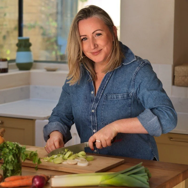 A woman with shoulder-length blonde hair, wearing a denim shirt, prepares vegetables in a kitchen, chopping onions on a wooden cutting board.