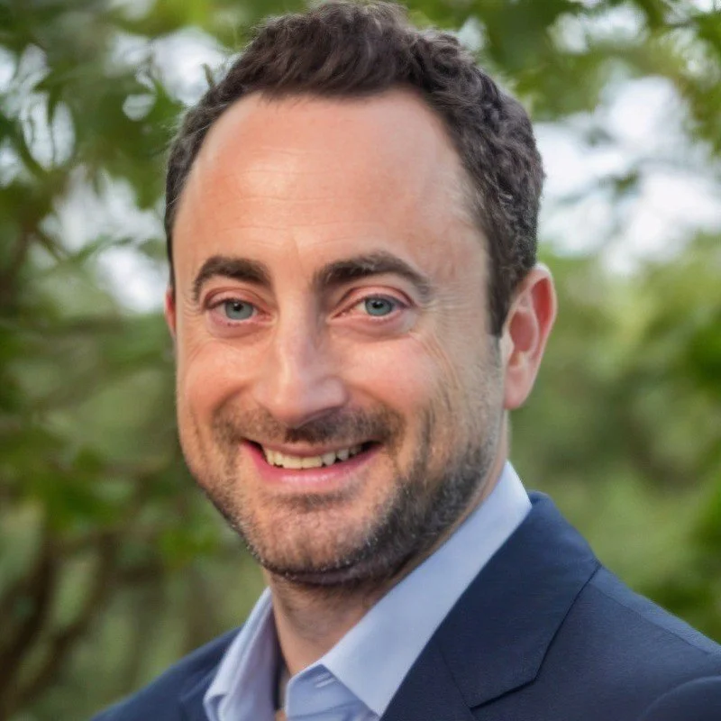 A portrait of a smiling man with short dark curly hair, light blue eyes, and a beard, wearing a navy blazer and light blue shirt, outdoors with blurred green foliage in the background.