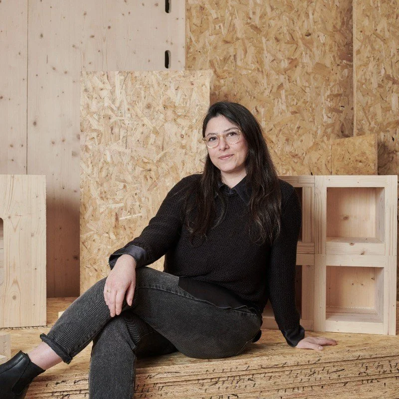 A woman sitting on a wooden platform in a workshop with plywood and OSB walls, wearing black jeans, a black sweater, and glasses.