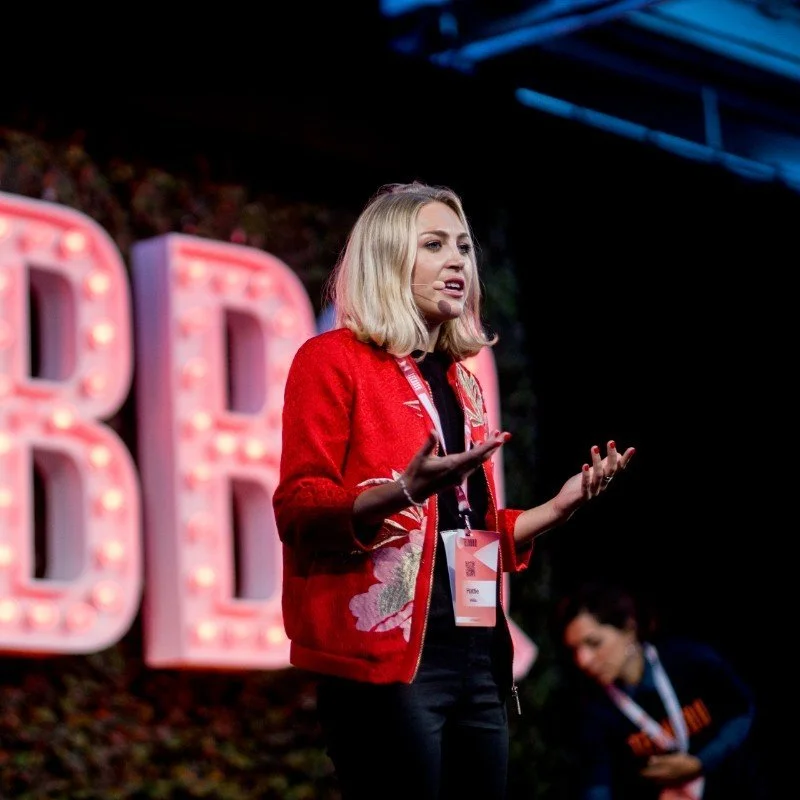 Woman with blonde hair speaking on stage at a conference or event, wearing a red jacket with floral embroidery, with a background of illuminated letters and a dark evening or indoor setting.