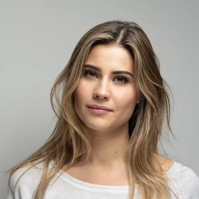 A young woman with shoulder-length, wavy blonde hair, wearing a white top, looking directly at the camera with a slight smile, against a plain gray background.
