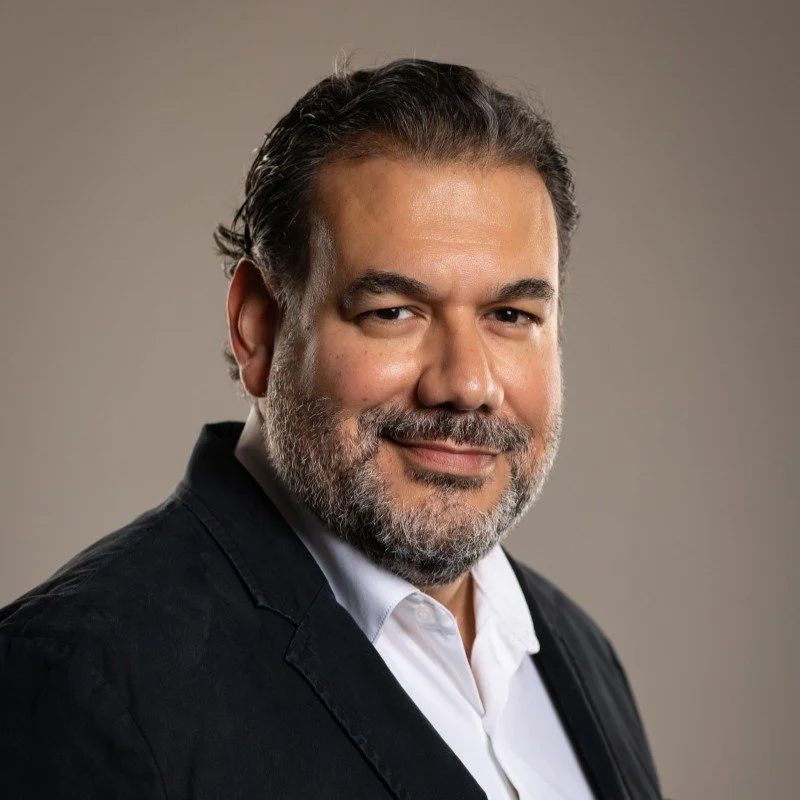 A man with dark hair and a beard, wearing a black blazer and white shirt, looking at the camera with a slight smile against a neutral background.
