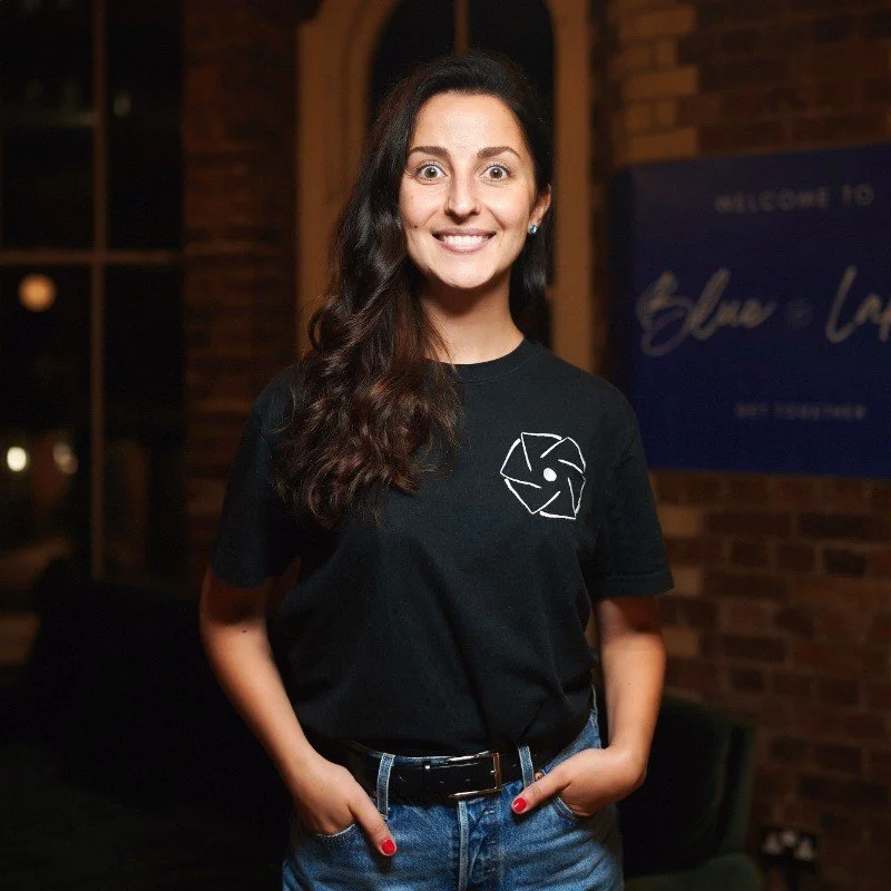 A woman with long dark hair, smiling and standing with her hands in her jeans pockets, wearing a black T-shirt with a white geometric lion logo, in front of a brick wall and window at night.