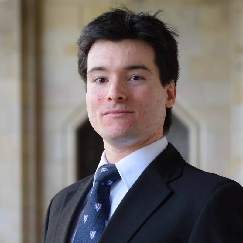 A young man in a black suit, white shirt, and a tie with small crest patterns, standing in front of a stone building, looking confidently at the camera.