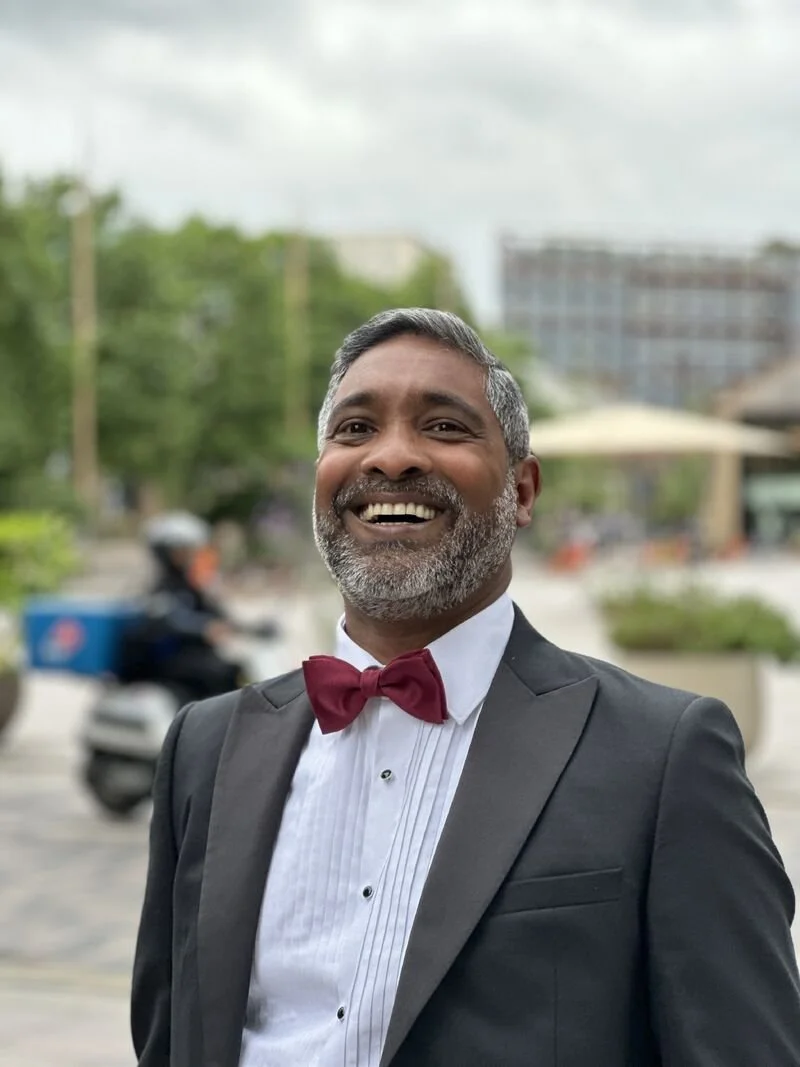 A smiling man in a tuxedo with a red bow tie, standing outdoors in an urban area with buildings, trees, and a motorcyclist in the background.