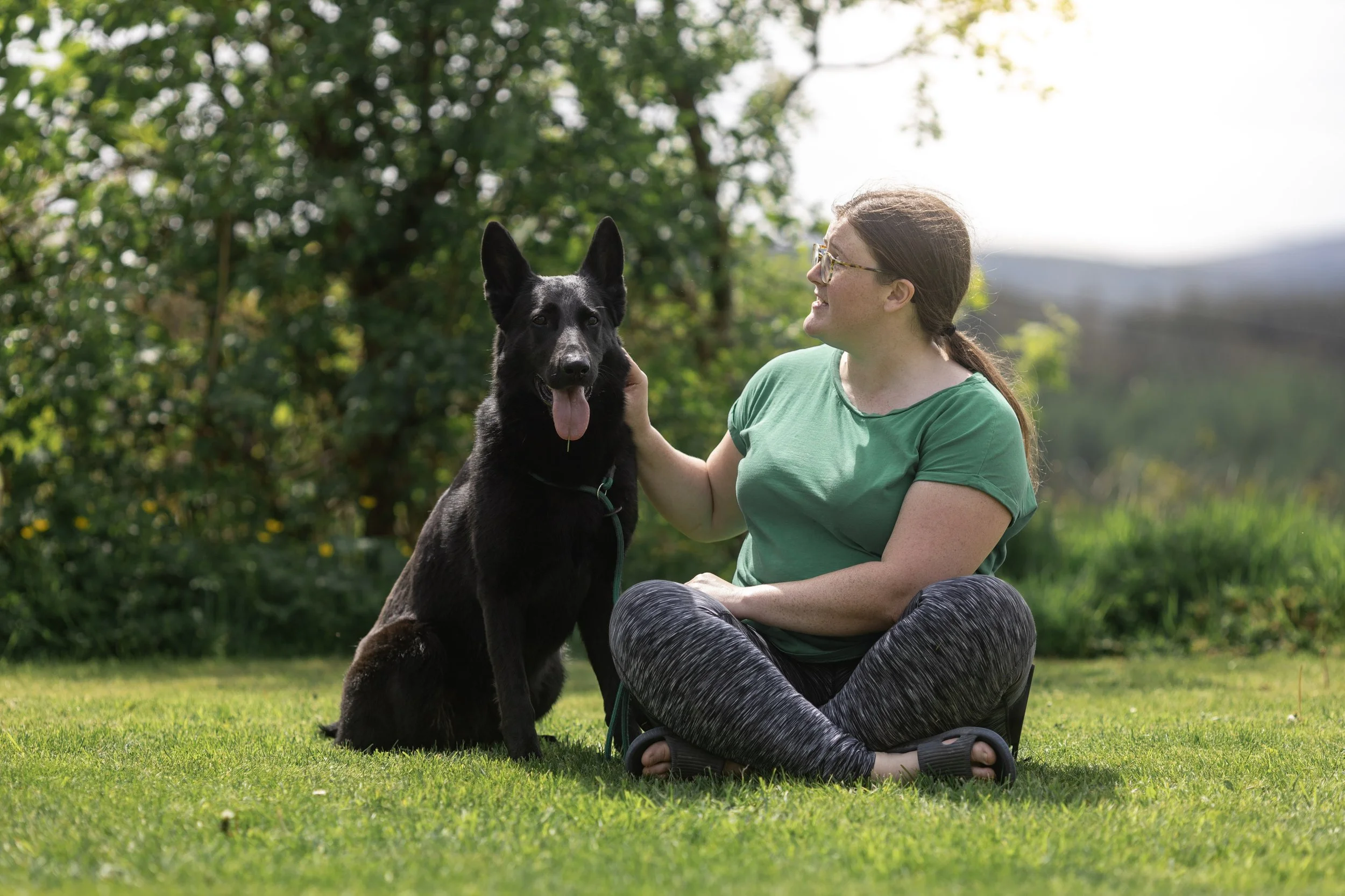A woman with brown hair, glasses, wearing a green t-shirt and patterned leggings, sitting cross-legged on grass, petting a black German Shepherd dog that is sitting beside her outdoors amidst greenery.