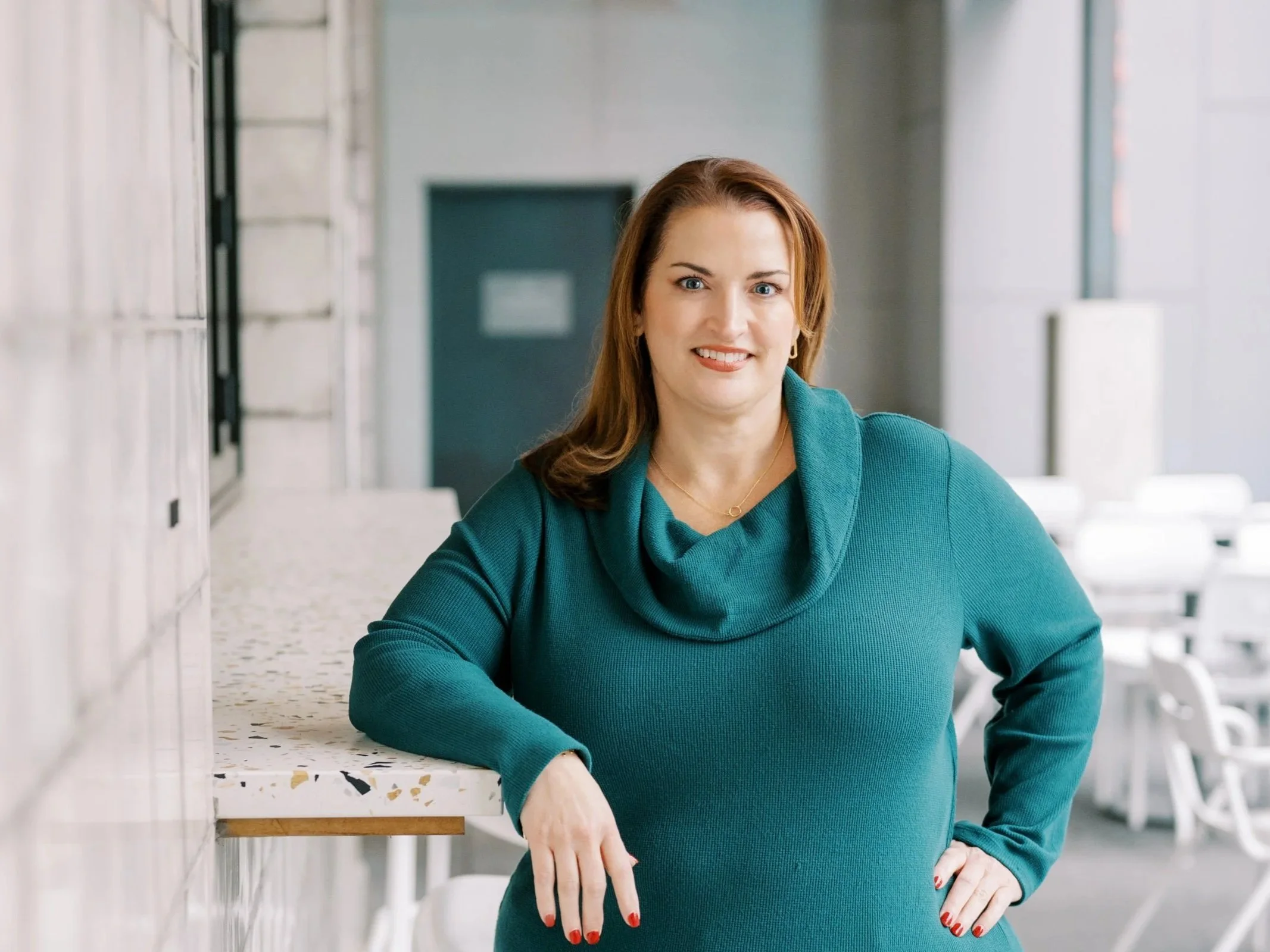 A woman with red hair, light skin, wearing a teal sweater, poses with her hand on her hip in a bright, modern space with large windows and white furniture.