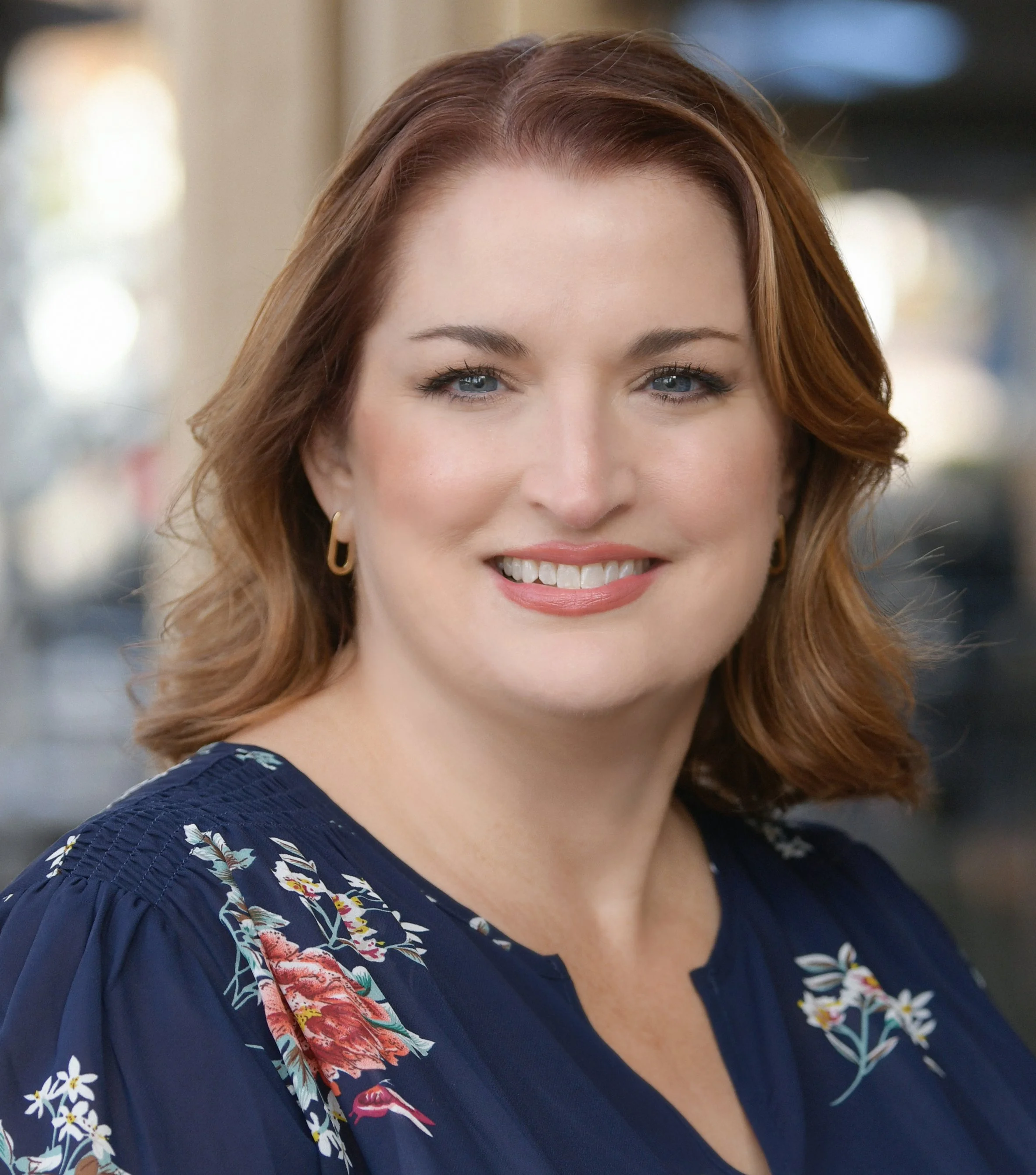 A woman with short, wavy auburn hair, wearing a dark blue floral blouse and gold earrings, smiling in an outdoor setting.