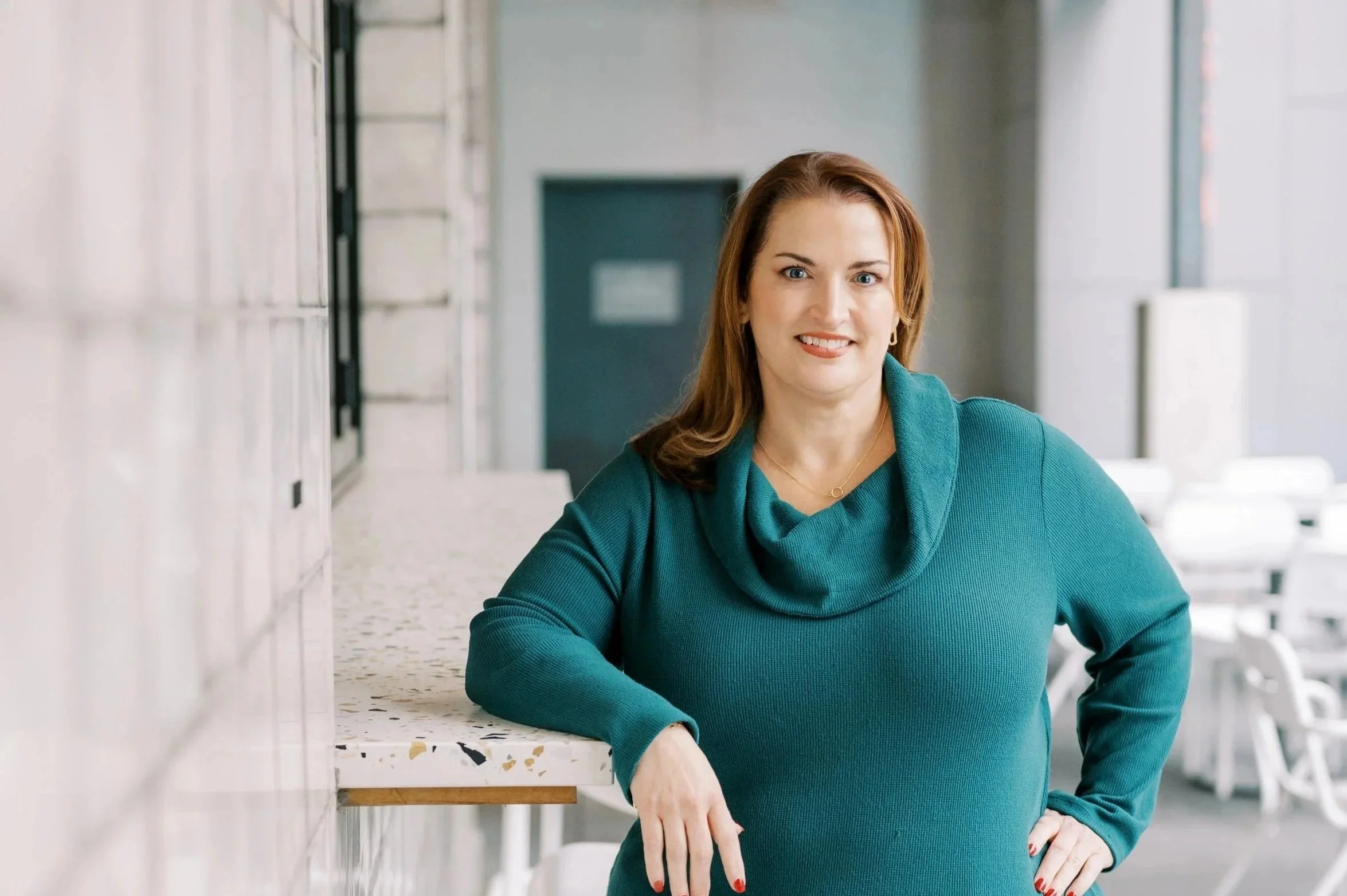 A woman with shoulder-length auburn hair, wearing a teal long-sleeved top with a cowl neck, smiling and leaning on a terrazzo counter in a modern, well-lit space with large windows and white chairs in the background.