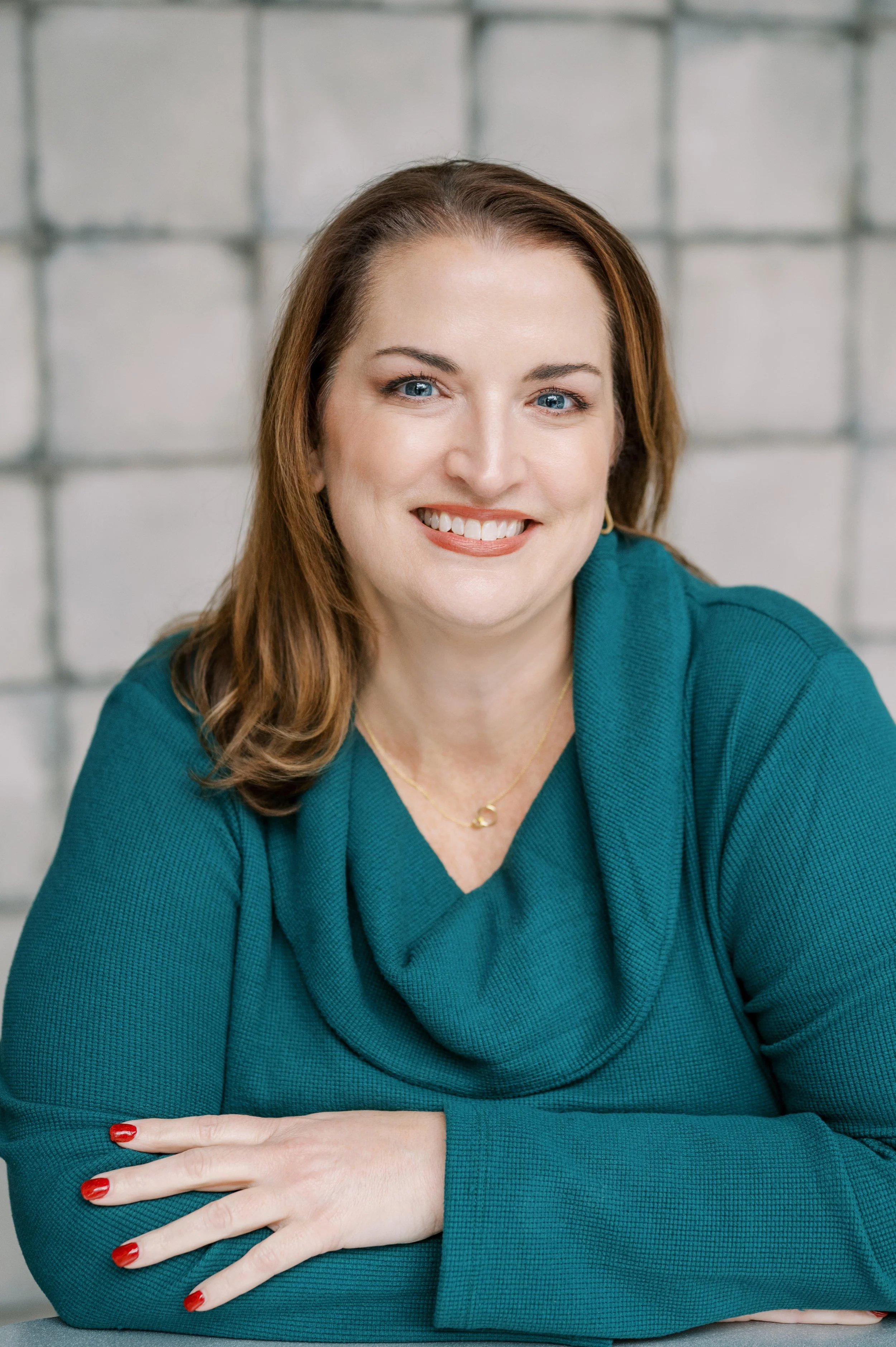 A woman with shoulder-length brown hair and blue eyes smiling, wearing a teal long-sleeve top and a gold necklace, sitting with her arms crossed in front of a gray brick wall.