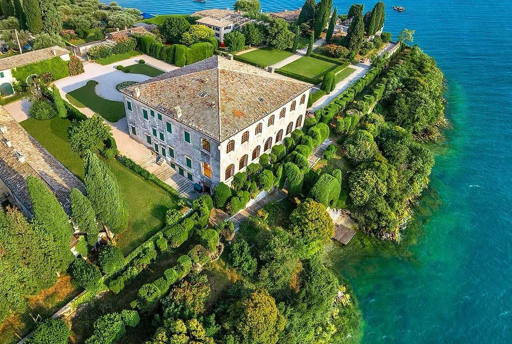 Aerial view of a large, historic villa surrounded by gardens and trees, adjacent to a lake with clear blue water.