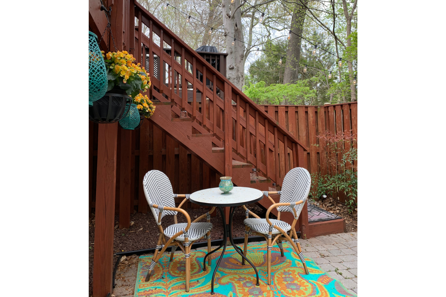 Small outdoor bistro seating area with string lights, patterned rug, and two chairs beneath a deck