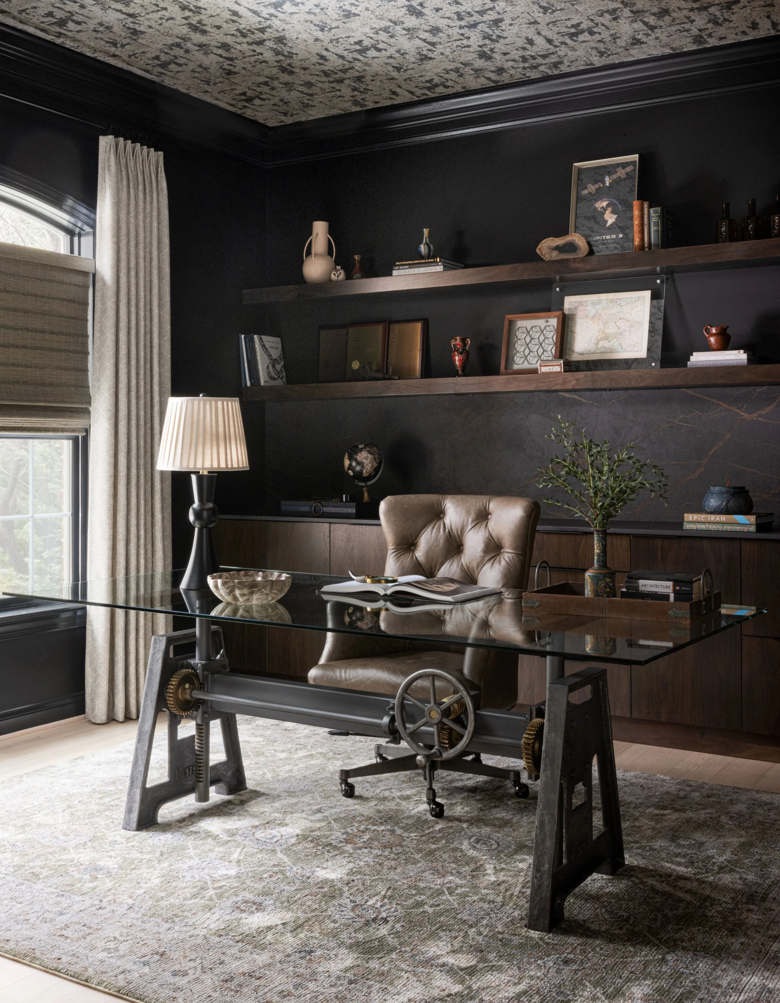 Luxury home office interior design by House of Shima featuring dramatic black walls, custom walnut built-ins, an industrial-style desk, vintage furnishings, layered lighting, and a patterned rug.